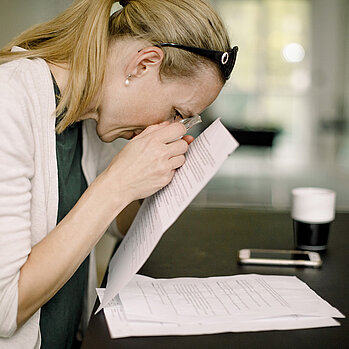 Frau mittleren Alters sitzt am Schreibtisch und liest mit Lupe ein Dokument